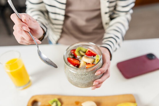 Close-up of a woman holding a jar of chia pudding topped with fresh fruit during a healthy breakfast, highlighting clean eating, nutrition and wellness with a smartphone nearby