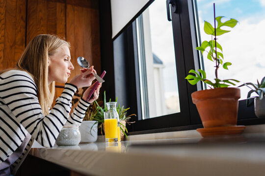 Peaceful morning scene with a young woman reading messages on her smartphone while enjoying fresh orange juice by the window. Calm home atmosphere, mindfulness, self-care and everyday happiness