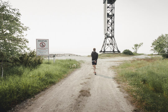 Saxby, Laane maakond, Estonia - 01 June 2025: View of a person walking along a dirt path towards the Saxby rannik coast, passing a warning sign and a tall metal beacon structure under a bright sky.