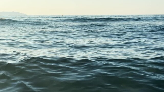Low angle seascape showing ocean waves advancing toward shore with undulating swells, scattered foam, bright whitecaps, reflective water, and natural coastal movement under daylight