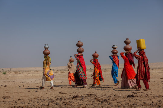 Yazman, Punjab, Pakistan - 19 March 2010: View of women in traditional colorful clothing walking across the arid Thar desert landscape while carrying water pots on their heads near Chanan Pir.