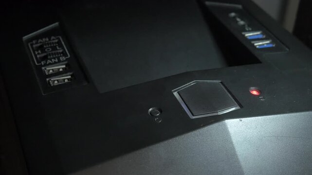 Close-up shot of a man's finger pressing the power button on a black computer case. The red LED indicator starts flashing to indicate that the computer is booting