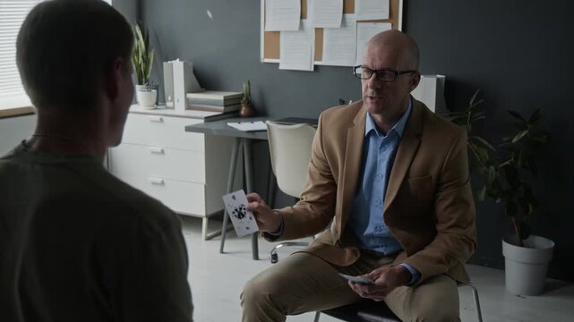 Middle-aged Caucasian male psychologist showing Rorschach inkblot cards to young adult Caucasian patient during therapy session. Footage for mental health and psychological services