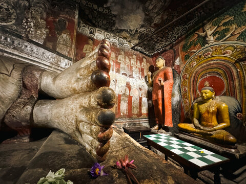 Sri Lanka - 20 April 2024: View of the reclining Buddha statue inside Dambulla Cave Temple, featuring ancient wall paintings, a seated Buddha figure, and lotus flower offerings.