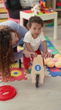 Woman guiding toddler's hands on wooden balance bike over colorful foam mats inside childcare building; care bonding joy.