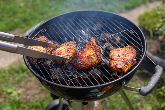Grilled pork chops cooking over charcoal outdoors. Metal tongs lift a browned pork chop as several seasoned chops cook on a smoky charcoal grill outdoors.