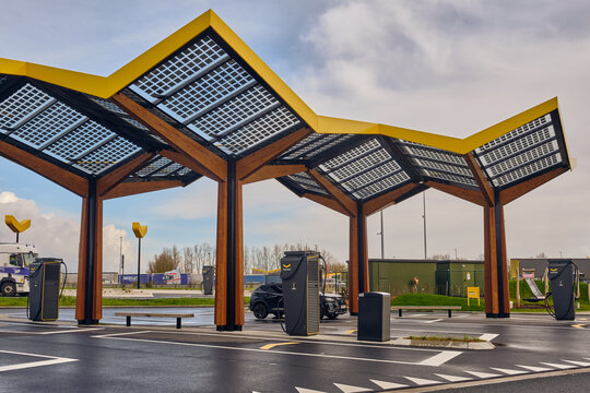 Gent, Belgium - 14 March 2026: View of a Fastned electric vehicle charging station featuring a distinctive yellow solar panel canopy and wooden support pillars.