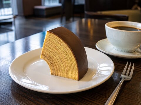 Feines St&uuml;ck Baumkuchen mit vielen sichtbaren Schichten und Schokoladen&uuml;berzug auf wei&szlig;em Teller, klassisch zu Kaffee serviert &ndash; deutsche Kuchenspezialit&auml;t.