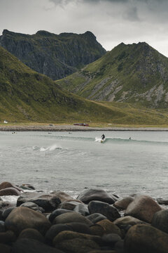 Bostad, Norway - 03 September 2025: View of a surfer riding a wave at Unstad Arctic Surf with rocky coastline and mountains in the background under a cloudy sky.