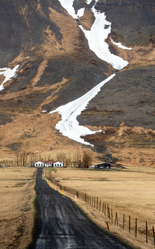 Winding country road leading to a remote farmhouse under a massive mountain with melting snow in Iceland.