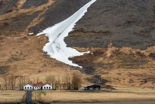 Iceland landscape of a paved country road with a wooden fence winding through a dry, golden grassy field towards a small white, red-roofed farmhouse situated at the foot of a giant, dark rocky mountai