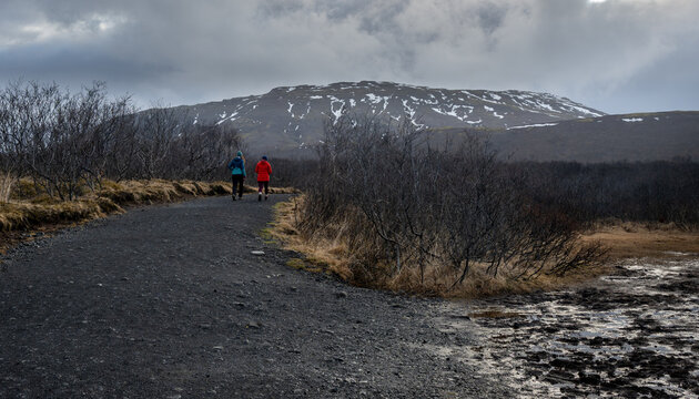 Two hikers walking gravel burrafoss path through a rugged landscape, with a snow capped mountain under a cloudy sky. Iceland Icelandic hiking trail