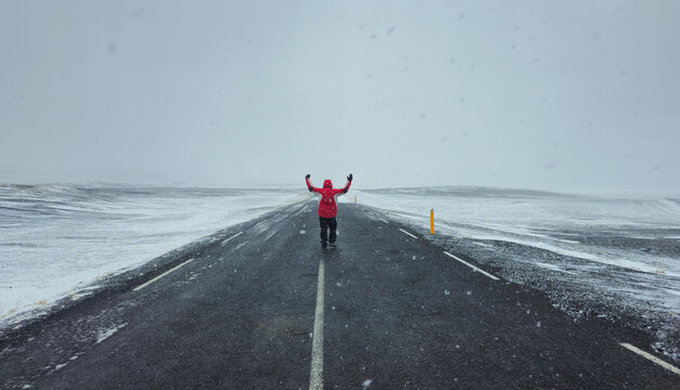 Lone Person in red jacket walking on a long, empty road during a snowstorm, holding arms wide, feeling freedom and adventure. Iceland stormy weather