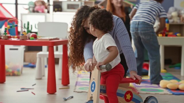 Woman holding toddler on wooden toy bike while supporting child in playroom building; nurturing play learning.