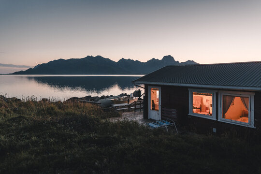 Holmstad, Nordland, Norway - 31 August 2025: View of a cozy wooden cabin with glowing interior lights by a calm fjord, with rugged mountains in the background under a twilight sky.