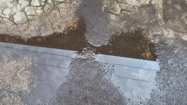 A high-angle shot of a puddle of rainwater on wet, cracked asphalt, reflecting the power lines and the sky above.