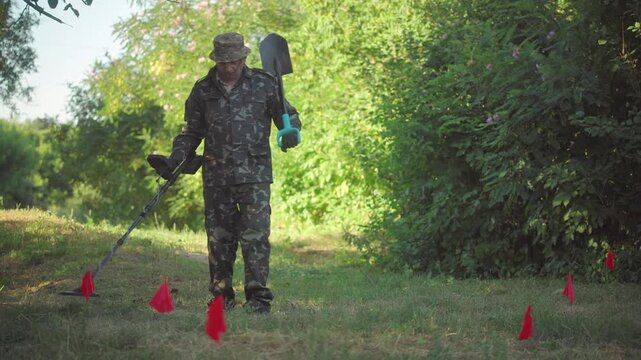 Male soldier in camouflage uniform uses a shovel and metal detector while surveying a grassy area marked with red flags, surrounded by dense green foliage and trees in a natural setting