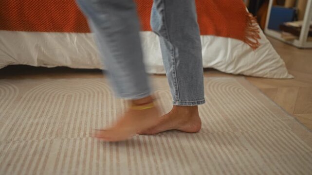 Woman barefoot stepping across striped rug by bed in studio, wearing cropped jeans and anklet with painted toes visible; calm morning routine.
