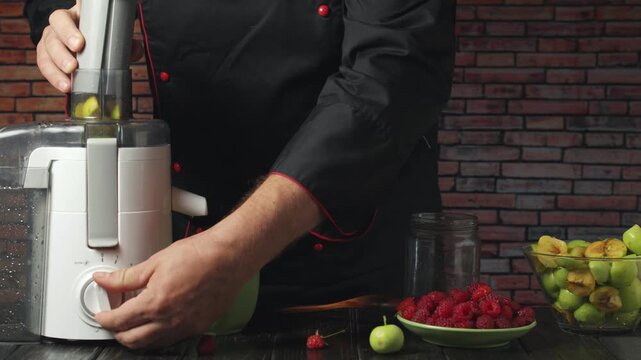 Chef in black uniform operates a juicer, pouring fresh juice into a green bowl while surrounded by raspberries, green apples, and jars on a wooden table against a brick wall backdrop