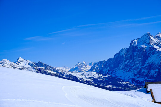 A stunning wide-angle winter landscape capturing a pristine, snow-covered mountain slope under a deep blue sky in the Jungfrau region. The photograph highlights a groomed snow trail curving gently acr