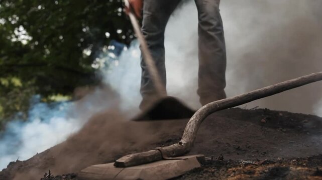 Low angle view of a worker tending a smoking charcoal mound with a traditional tool. Artisanal charcoal production, rural manual labor and traditional craft concept.