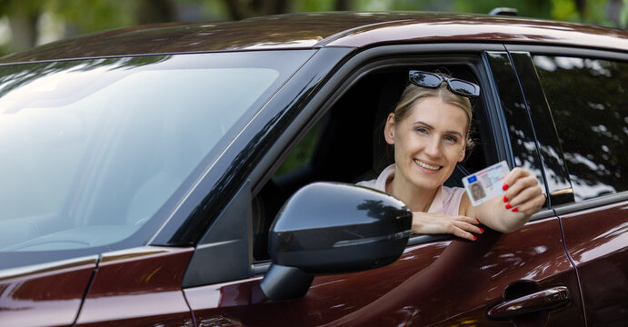Smiling woman sitting in the car and showing her new driving license out of window to the camera
