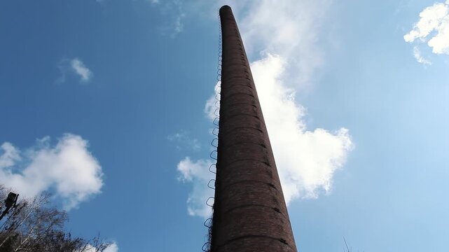 Low angle view of an old tall brick industrial chimney with a metal ladder against a blue sky with clouds. Vintage factory and abandoned industry concept.