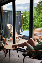 Obraz premium Woman relaxing indoors with legs on table, looking through large window at mountain landscape, cozy modern cabin interior with terrace, outdoor hot tub and lounge chairs