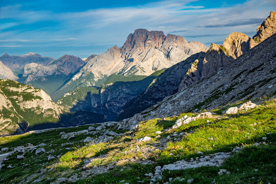 Expansive mountain landscape featuring steep rocky slopes, green summer grass, and distant peaks with sunlight illuminating their rugged forms under a blue sky in the Italian Dolomites