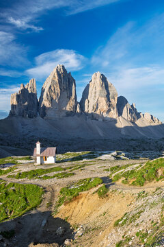 Chapel standing on a hillside with the iconic Tre Cime di Lavaredo peaks rising majestically under a clear blue sky in the rugged Dolomite Alps of Italy