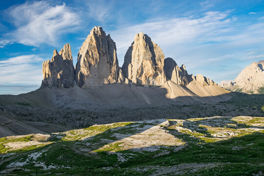 Tre Cime di Lavaredo. Iconic jagged peaks in the Italian Dolomites. Bathed in golden sunlight. Showcasing a stunning alpine summer landscape with partial shadows on the rugged terrain and green grass