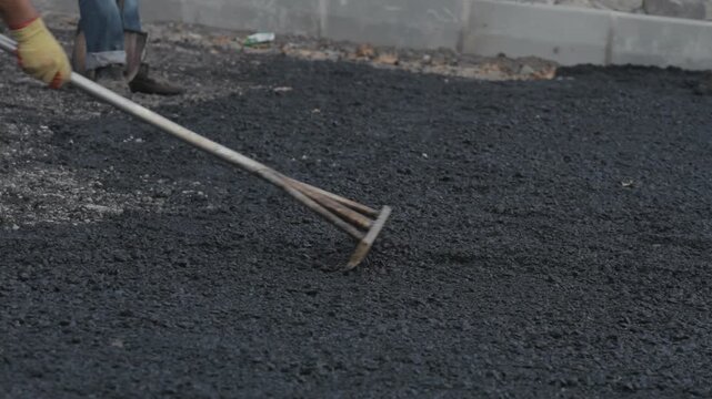 Close-up of a construction worker using a rake to level and spread fresh black asphalt. Manual labor, road building, and infrastructure repair concept.