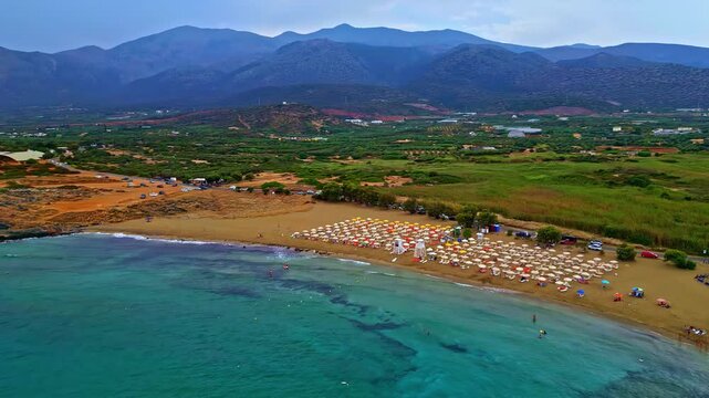 Aerial view of Crete beach featuring rows of umbrellas on the sand, turquoise sea, and rolling mountains in the background, Greece.
