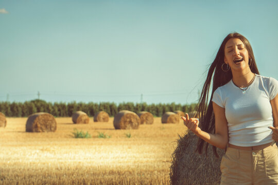 A vintage snapshot captures a teenage girl celebrating freedom in a sun-drenched hayfield. She's dancing with pure joy, lost in the music and the natural beauty of the summer countryside. Soft lens fl