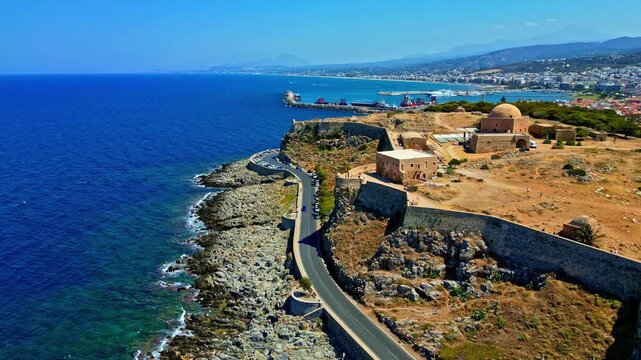 Aerial view of the historic Fortezza of Rethymno, featuring ancient stone walls, a prominent dome, and a coastal road winding along the rocky Mediterranean shoreline in Crete, Greece.