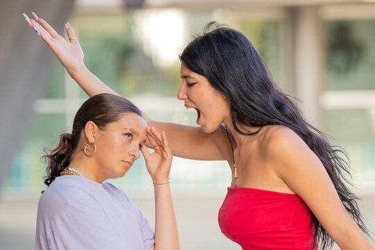 Young hispanic woman in red top yelling and gesturing at unbothered friend outdoors. Conflict, drama, and attitude concept