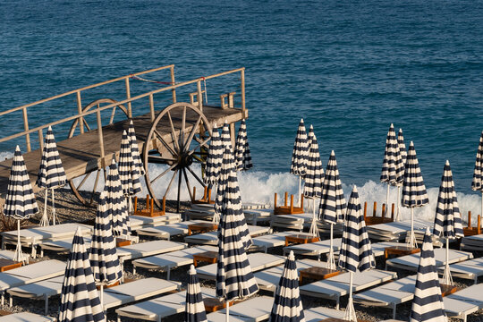 Striped beach umbrellas and folded sunbeds on a pebble beach with a wooden pier in Nice Cote d Azur France