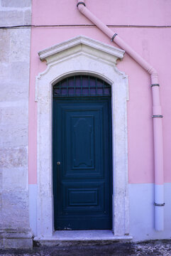 A dark green wooden door with a decorative carved stone frame stands set into a pastel pink building facade in Lisbon, Portugal