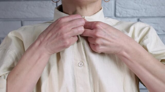 Woman unbuttoning her beige cotton shirt. Close-up view of an unrecognizable woman's hands unbuttoning her classic beige cotton shirt against a gray brick wall