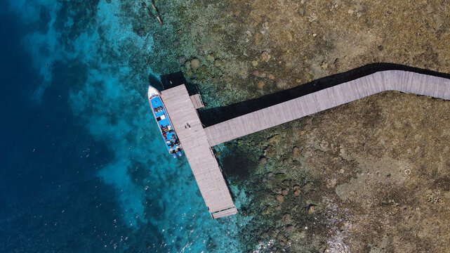 Aerial view of a boat docked at a wooden pier over vibrant coral reefs in the clear turquoise waters of Raja Ampat, Southwest Papua, Indonesia.