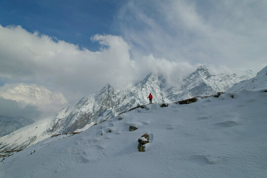 Aerial view of a hiker in a red jacket standing on a snow-covered mountain ridge with the majestic Himalayan peaks and clouds in the background in Nepal.