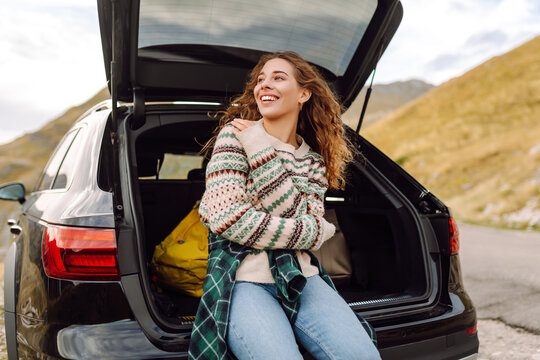 A young woman in a cozy sweater sits in the trunk of a car, enjoying the mountain views. Wanderlust. The concept of nature, transportation, and enjoyment.