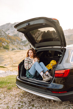 A young woman in a cozy sweater sits in the trunk of a car, enjoying the mountain views. Wanderlust. The concept of nature, transportation, and enjoyment.