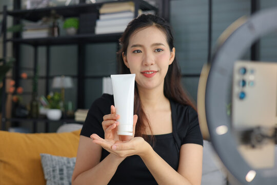 Asian woman filming beauty vlog tutorial with ring light, reviewing cosmetics cream moisturizer or lotion.