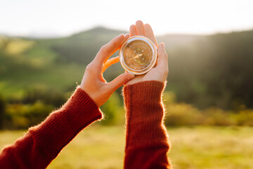 A rear view of a hiker in a hat holding a navigational compass. A woman stands atop a green hill, holding a compass, at sunset. Concept of direction, adventure, and nature. © maxbelchenko