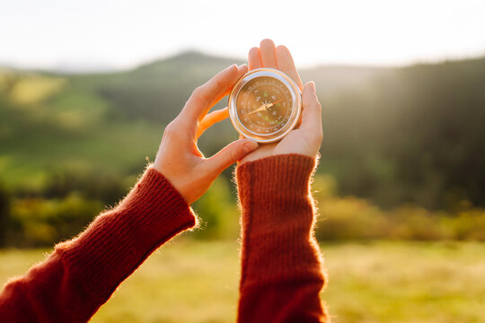 A rear view of a hiker in a hat holding a navigational compass. A woman stands atop a green hill, holding a compass, at sunset. Concept of direction, adventure, and nature.
