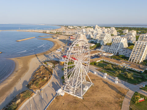 Aerial view of a large Ferris wheel situated on the sandy beach near the distinctive pyramid-shaped architecture of La Grande-Motte, Occitanie, France.
