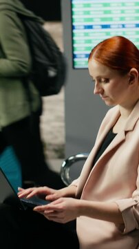 Vertical video: A businesswoman focuses on laptop work near an airport gate, managing responsibilities before an international flight while traveling abroad as a general manager for corporate travel.