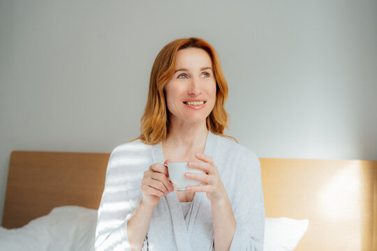 Relaxed smiling middle aged woman with cup of coffee relaxing in bed in morning sunlight. Hot drink for wake up and energy boost. Well being morning routine and daydreaming mood. Good morning habits.