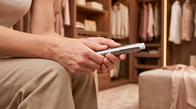 Close-up of a woman's hands holding a medical injector pen while sitting in a modern walk-in closet, preparing for a self-injection of medication